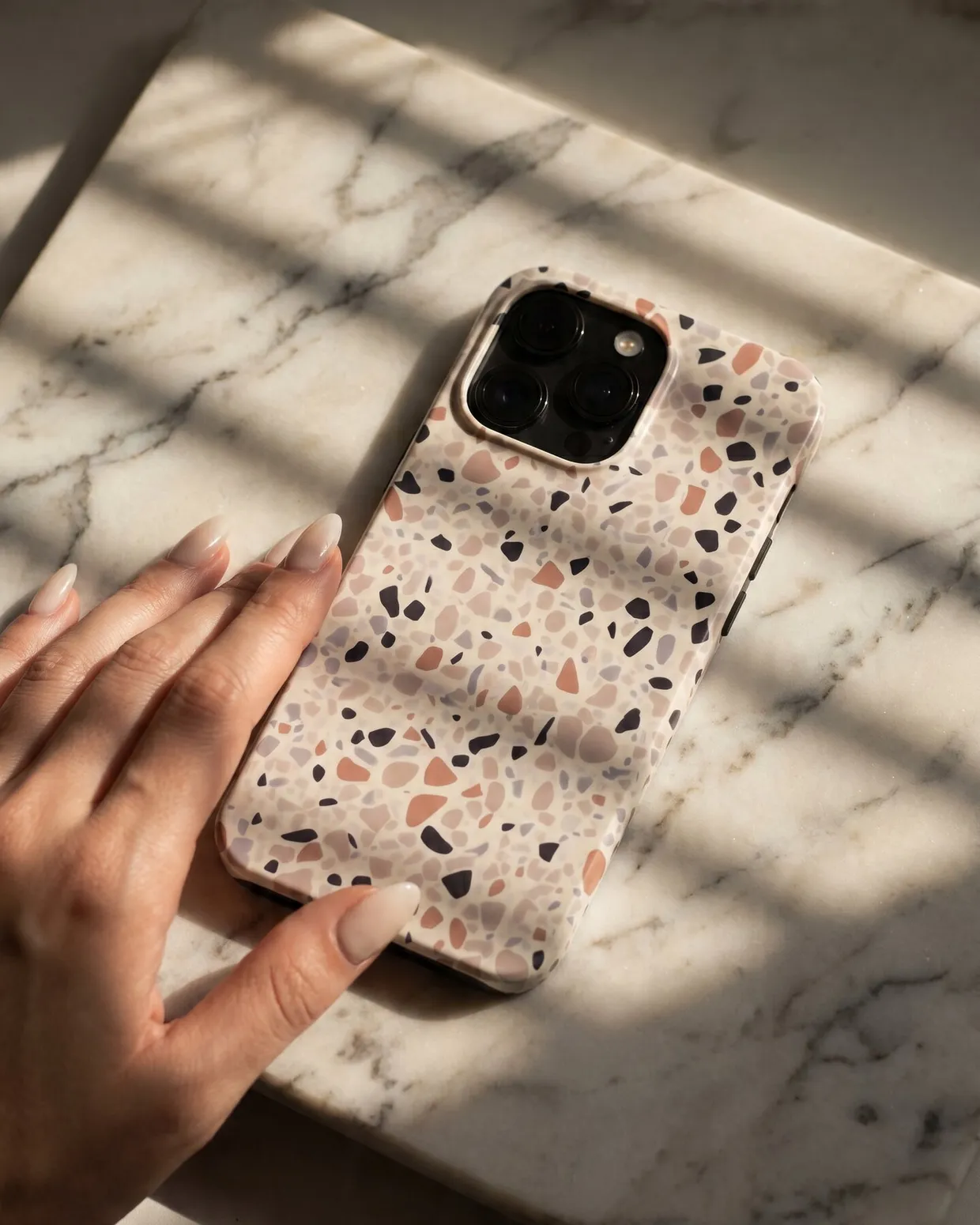 Terrazzo pattern iPhone case resting on a marble table, touched by a woman's hand